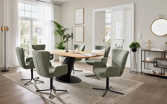 Helles Esszimmer mit modernem Esstisch und sechs grünen Stühlen. Große Fenster lassen viel Licht herein, während Pflanzen das Ambiente betonen.