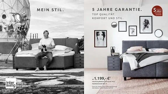 Ein Mann sitzt lächelnd auf einem Sofa am Strand, mit einem Bett im Hintergrund und Wellen im Meer. Er genießt die ruhige Atmosphäre.