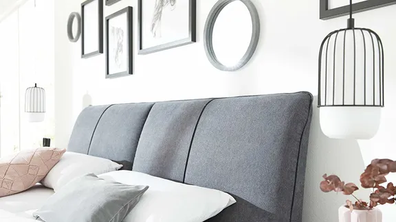 Detailaufnahme eines dunkelgrauen Polsterbetts mit weißer Bettwäsche und grauer Tagesdecke in einem modernen, hellen Schlafzimmer.