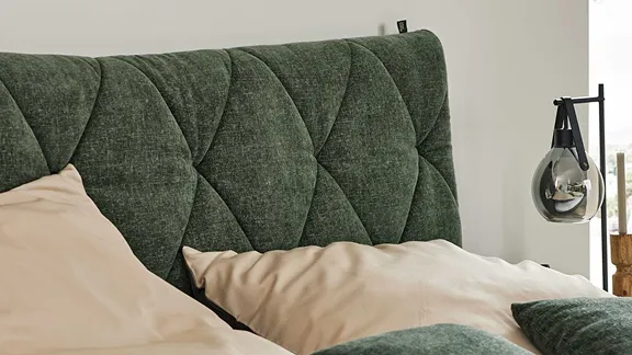 Nahaufnahme eines grünen Polsterkopfteils mit Rautensteppung, davor Kissen und eine moderne Glaslampe.