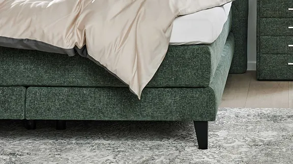 Detailansicht des Fußteils eines grünen Polsterbetts mit schwarzen Füßen, heller Bettwäsche und grauem Teppich.